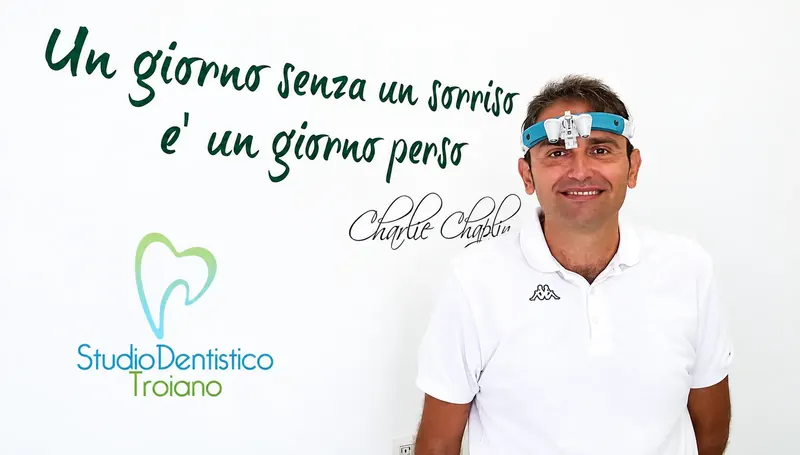 Il Dott. Amedeo Troiano, sorridente, in polo bianca e con gli occhiali ingranditori, davanti alla parete dello studio con la frase «Un giorno senza un sorriso è un giorno perso»
